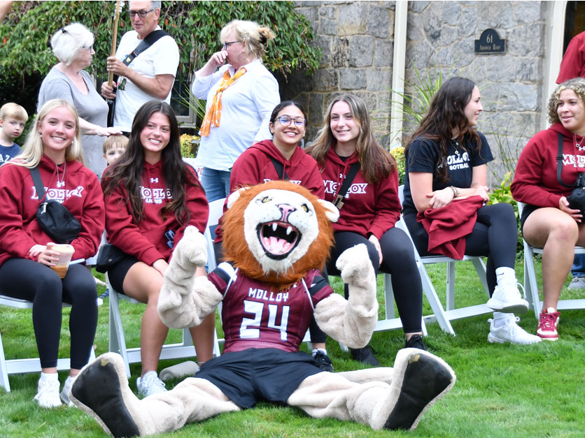 Molloy University students wearing Molloy Lions hoodies and shirts sit in chairs next to Molloy University Lion mascot