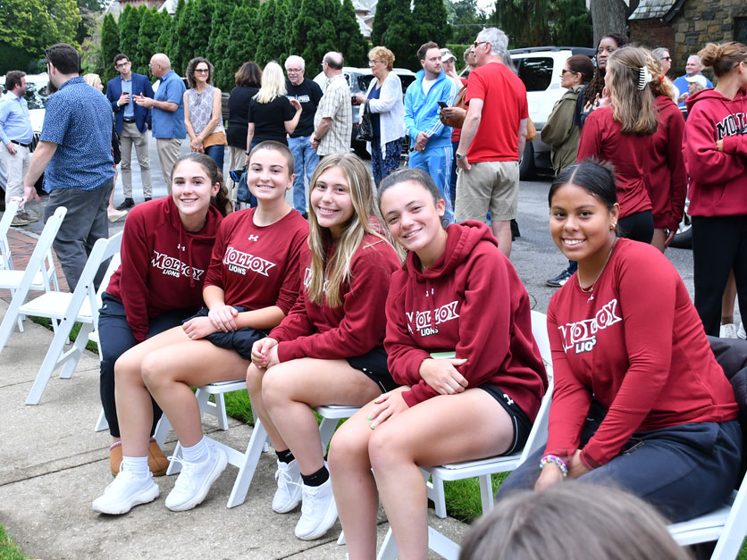 Molloy University students smiling, wearing red Molloy Lions hoodies sit on white chairs on Community Day